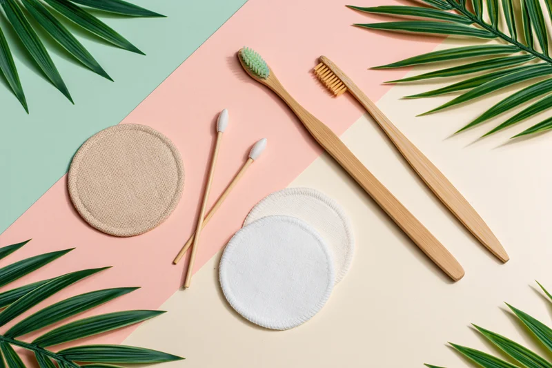 Bamboo toothbrushes and reusable cotton pads arranged in flat lay with palm leaves on pastel background
