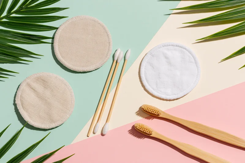Reusable cotton pads, bamboo toothbrushes, and cotton swabs arranged in a flat lay with palm leaves on a pastel background