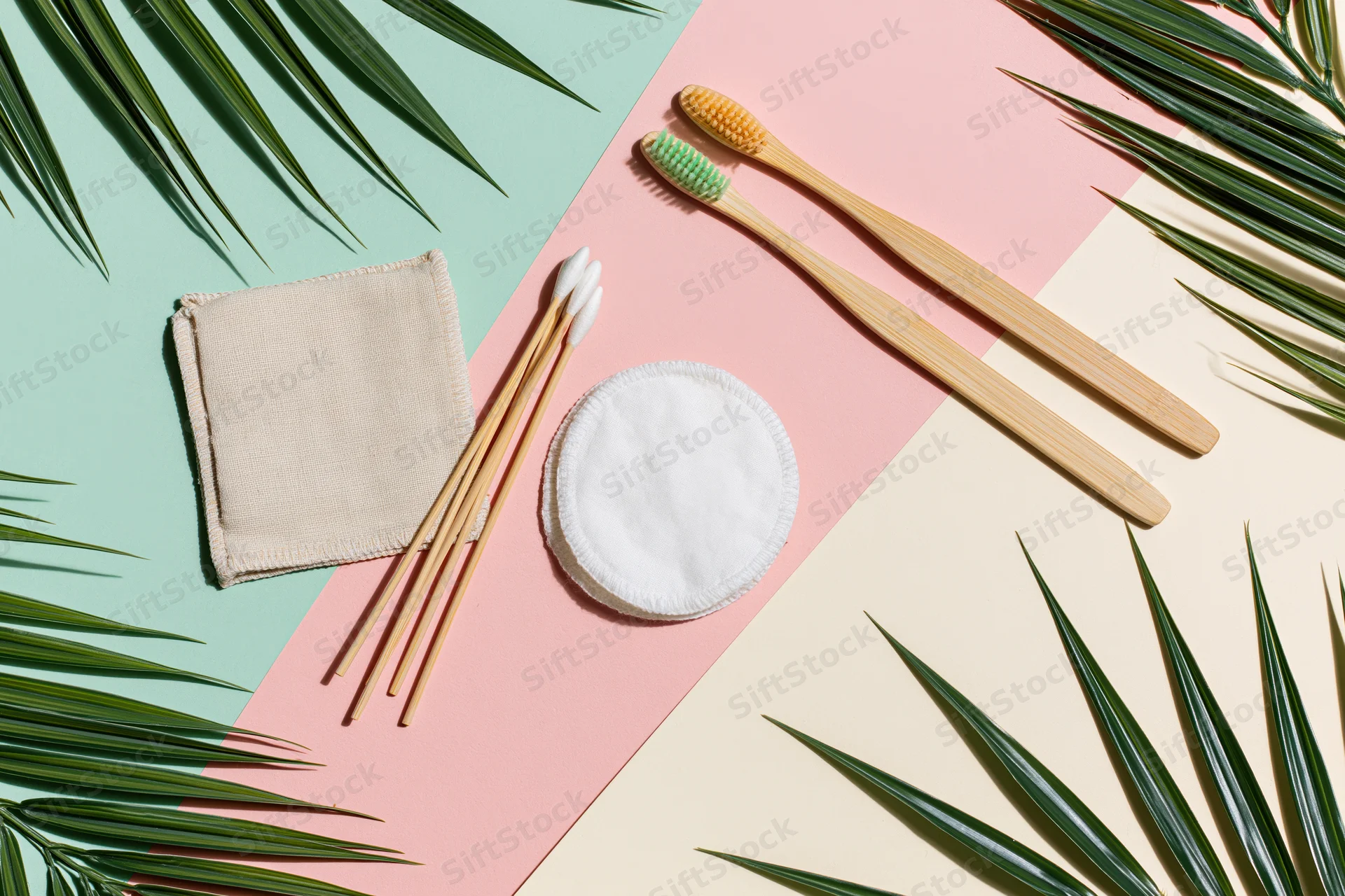 Bamboo toothbrushes and reusable cotton pads arranged flat on pastel background with palm leaves