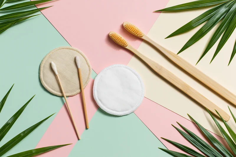 Bamboo toothbrushes and reusable cotton pads arranged in flat lay with palm leaves on pastel background