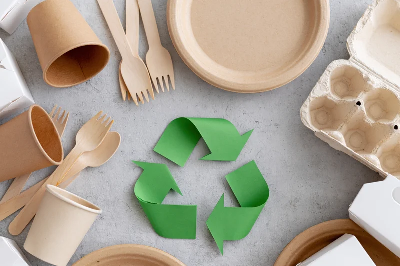 Eco-friendly disposable tableware arranged in flat lay on gray surface with recycling symbol in center