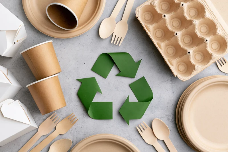 Eco-friendly disposable tableware arranged in flat lay around recycling symbol on gray surface
