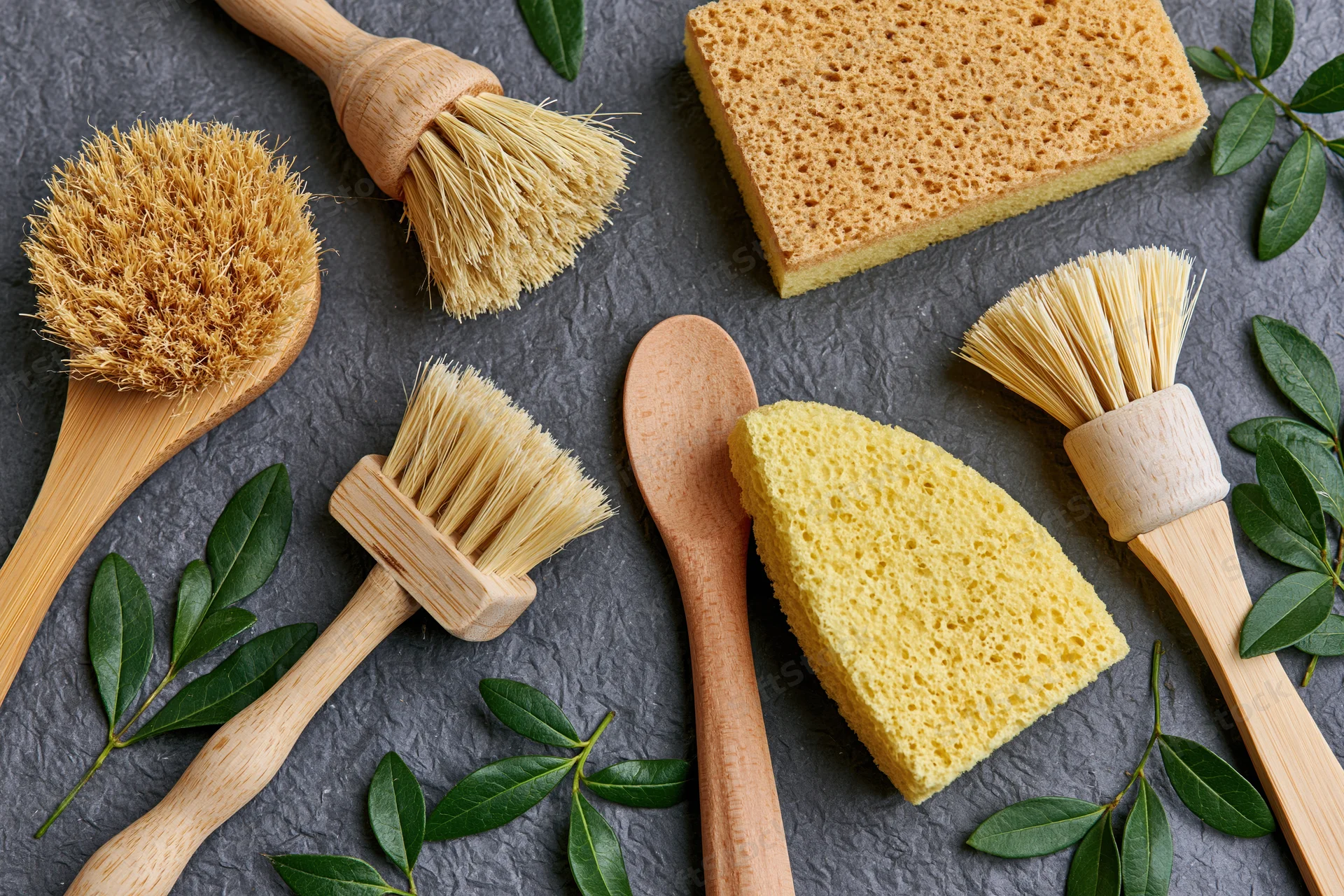 Wooden dish brushes and yellow sponges arranged flat on dark stone surface with green leaves