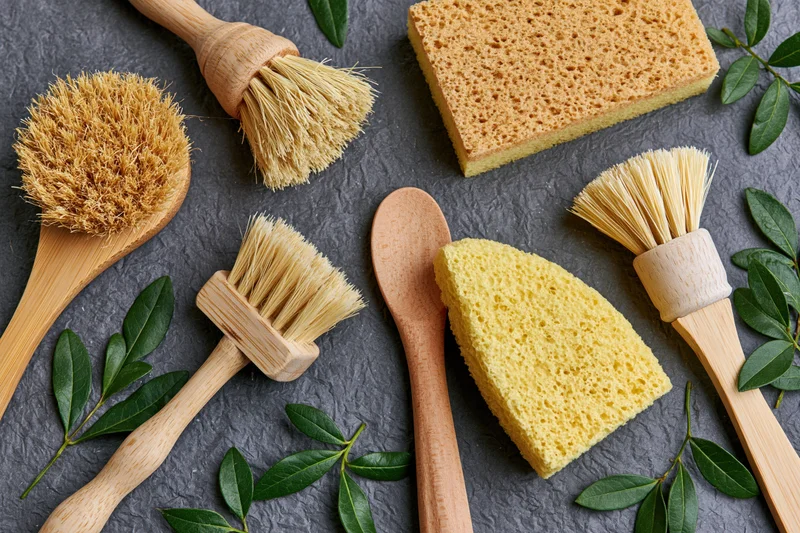 Wooden dish brushes and yellow sponges arranged flat on dark stone surface with green leaves