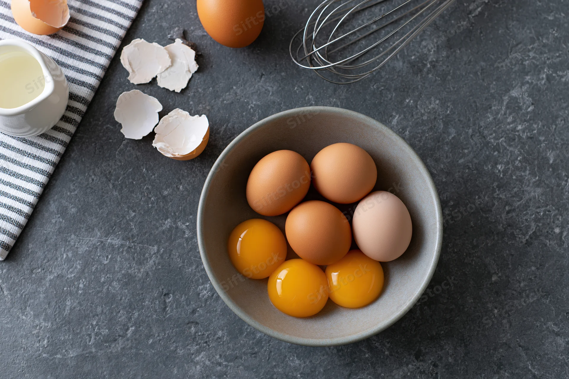 Five raw egg yolks and whole eggs in a matte ceramic bowl on a dark countertop with cracked shells, milk, whisk, and striped napkin nearby