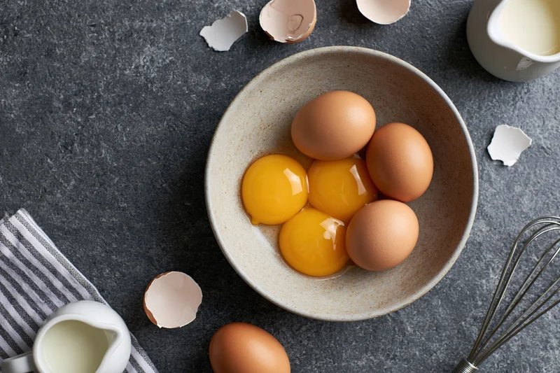 Five raw egg yolks and whole eggs in a matte ceramic bowl arranged in a flat lay on a dark countertop with milk pitchers, eggshells, and a whisk nearby