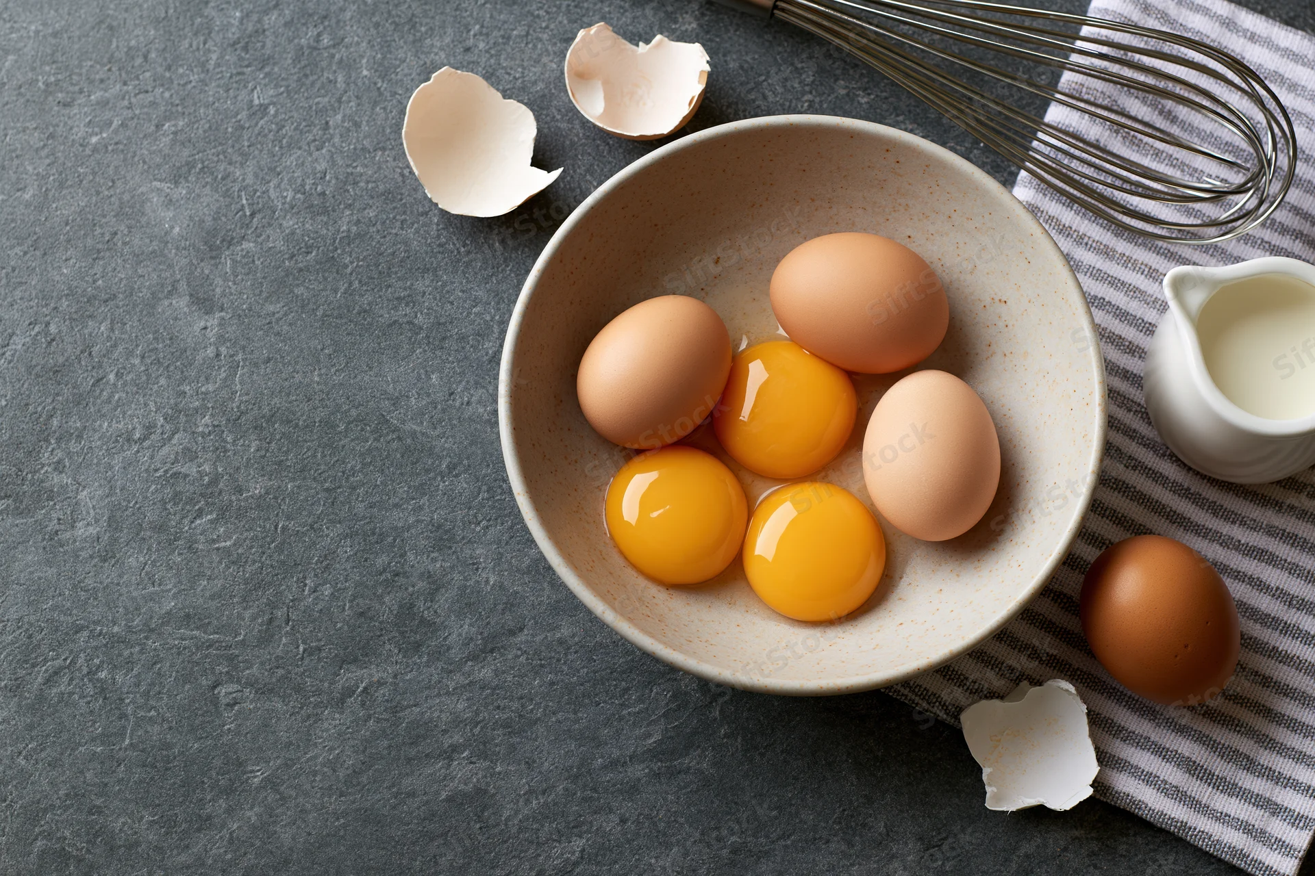 Five raw egg yolks and whole eggs in a matte ceramic bowl on a dark countertop with cracked shells, whisk, cloth, and milk pitcher arranged in flat lay