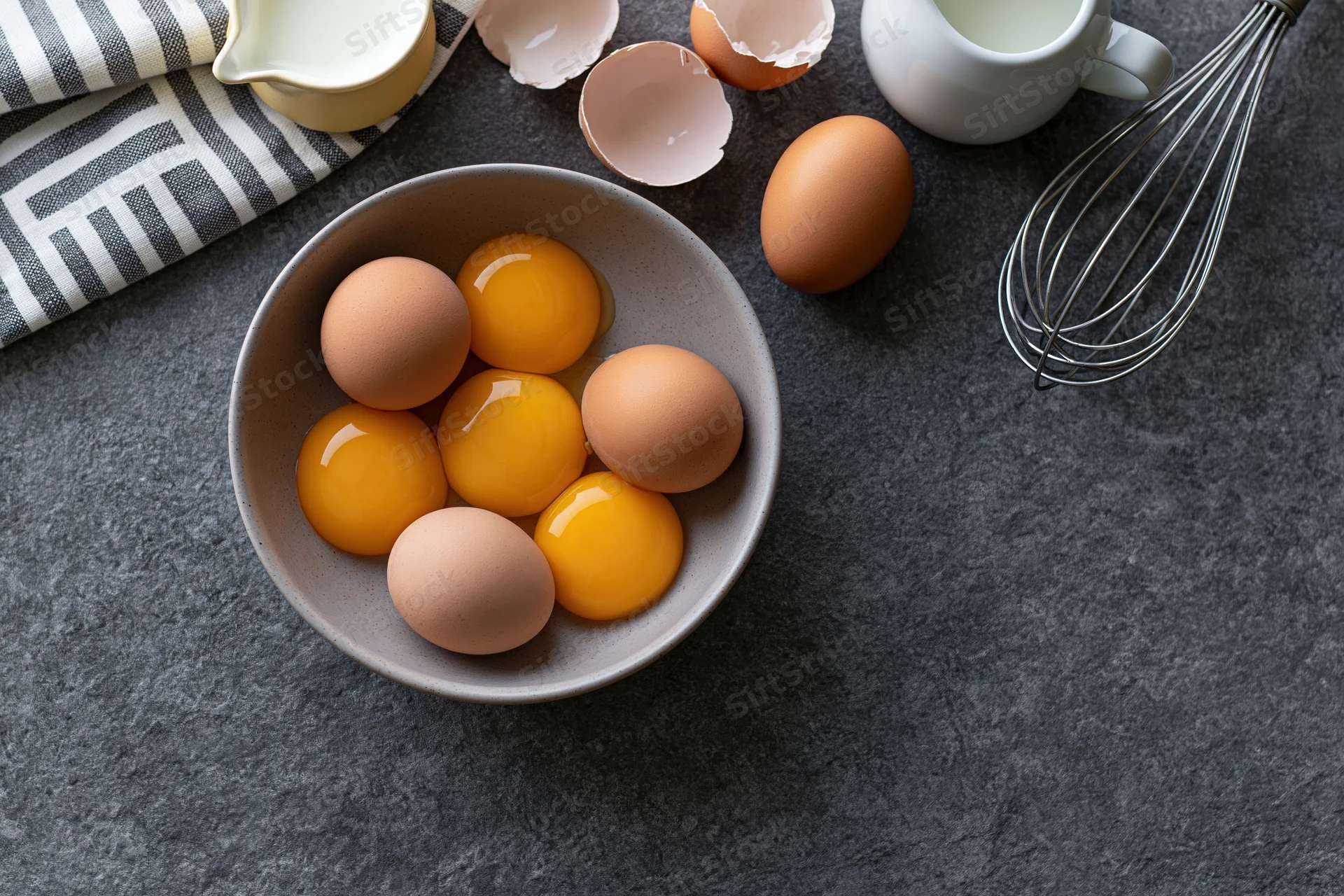 Five raw egg yolks and whole eggs in a matte ceramic bowl arranged on a dark countertop with milk pitchers and a whisk nearby