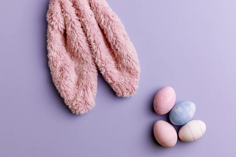 Fluffy pink plush bunny ears and four pastel decorated eggs arranged on a lavender background in a flat lay composition