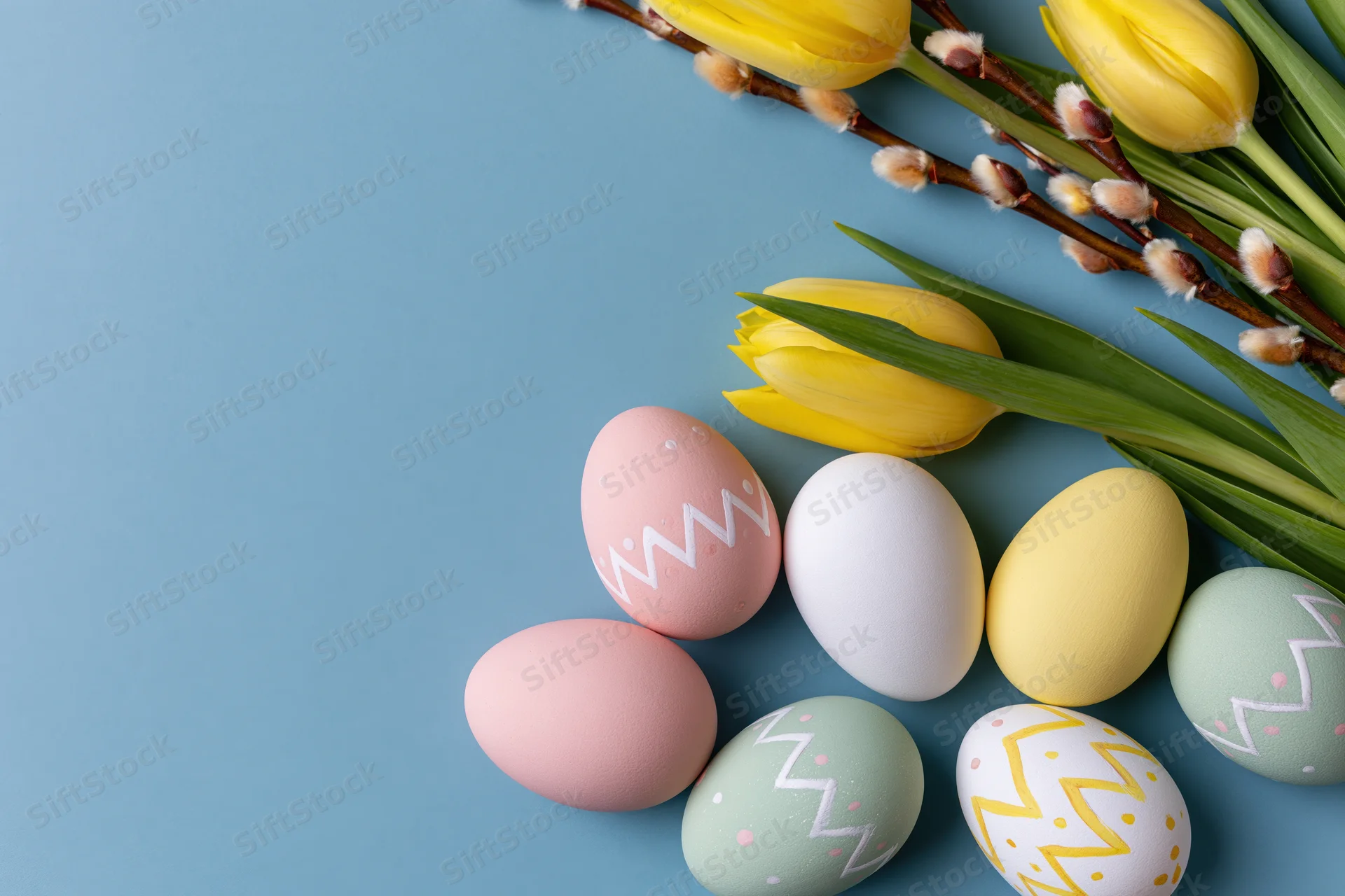 Hand-painted easter eggs and yellow tulips arranged in a flat lay on a blue background with copyspace