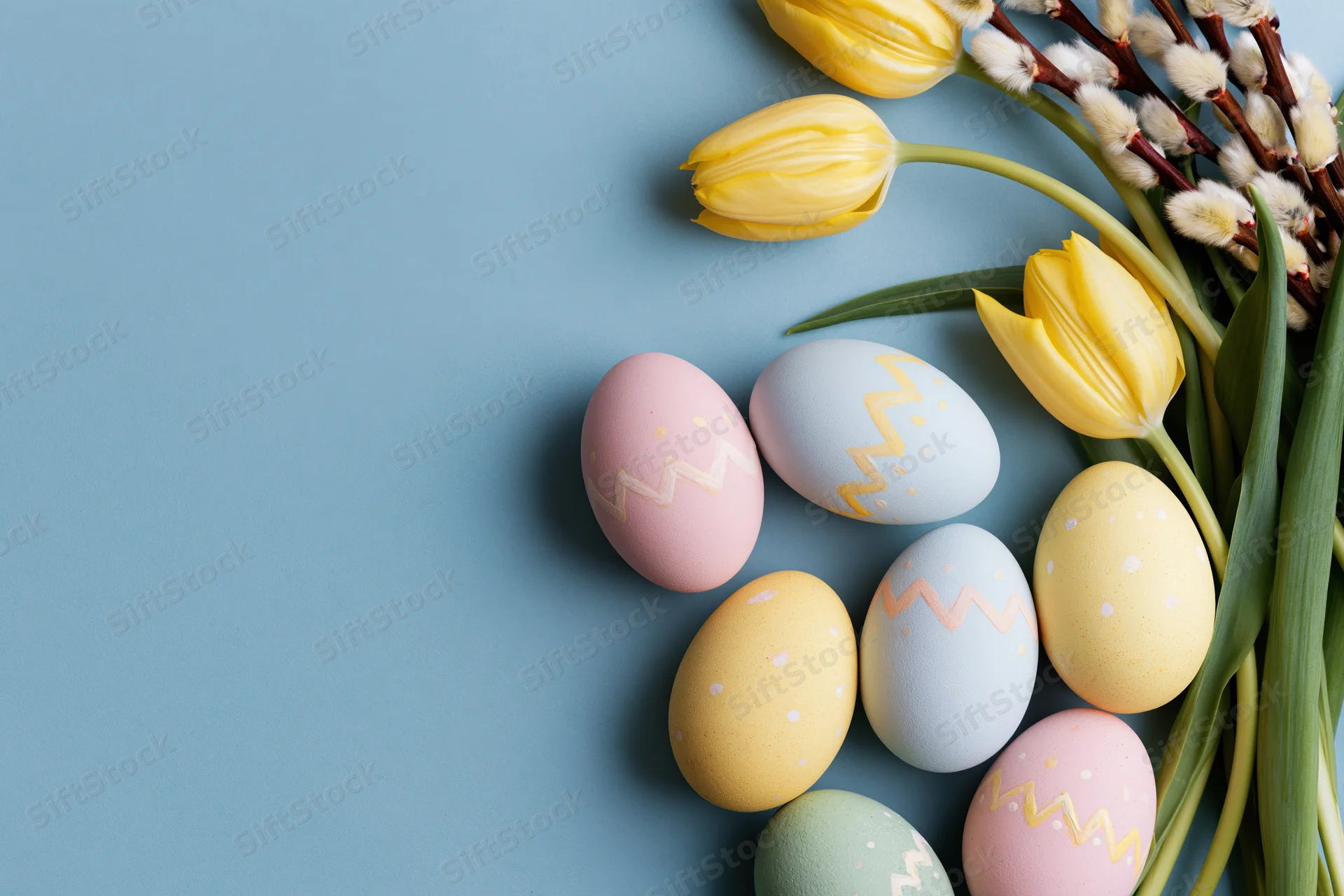 Hand-painted pastel Easter eggs and yellow tulips with willow arranged in flat lay on blue background