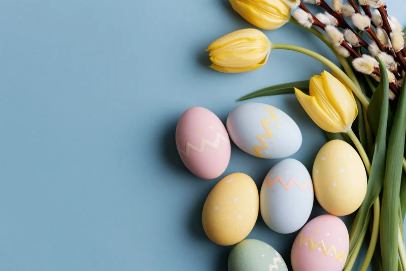 Hand-painted pastel Easter eggs and yellow tulips with willow arranged in flat lay on blue background