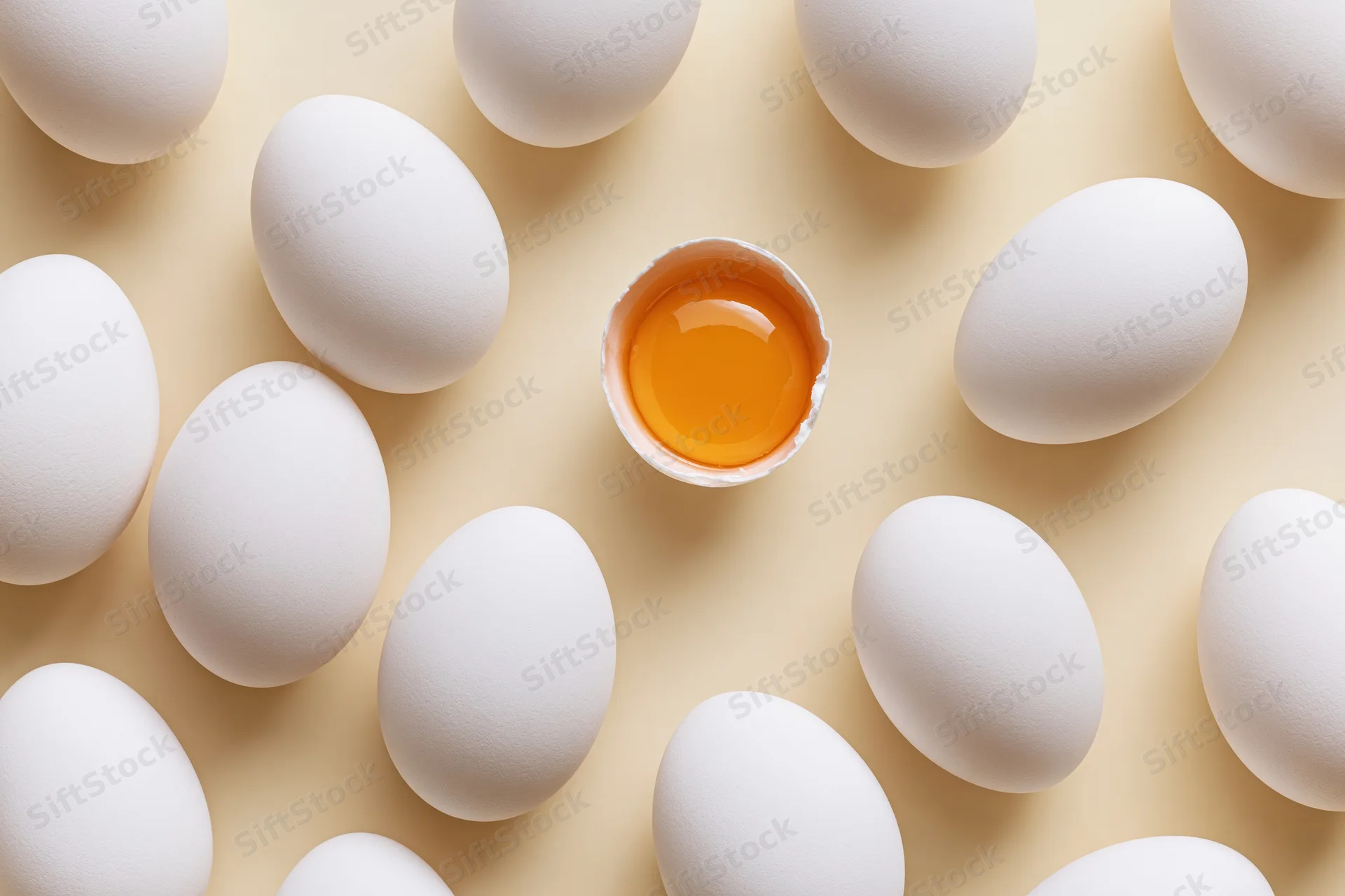 Multiple whole white chicken eggs arranged in flat lay with one broken egg showing yolk in center on beige surface