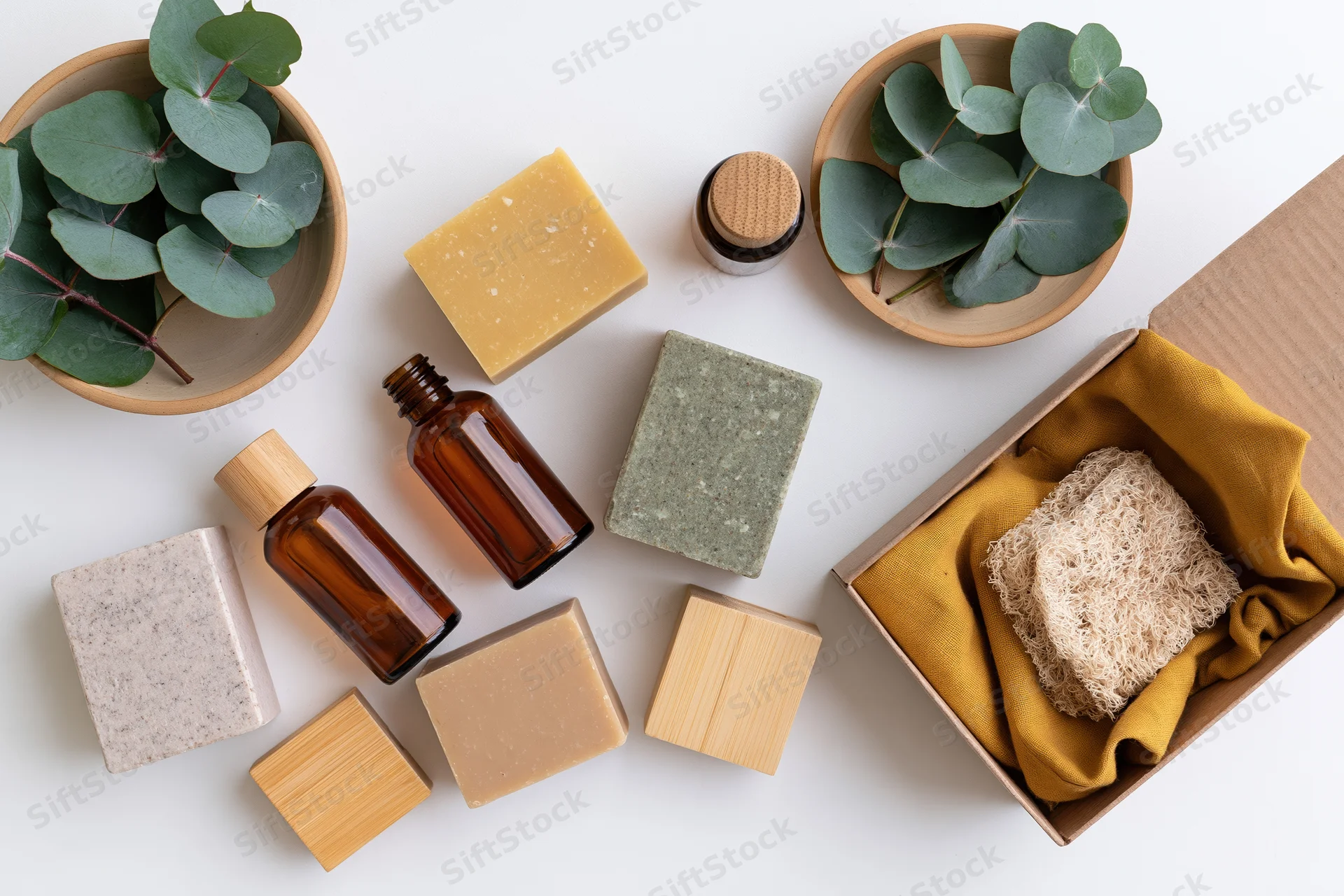 Natural skincare products arranged in flat lay on white surface with eucalyptus leaves and bamboo accessories