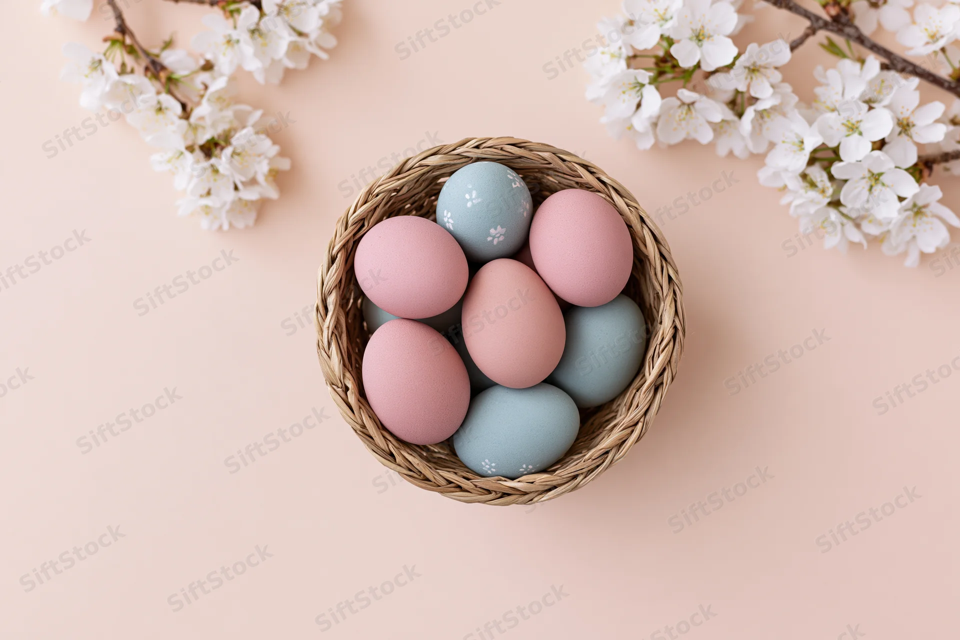 Pastel-painted easter eggs in a small woven basket with cherry blossoms on a soft peach background