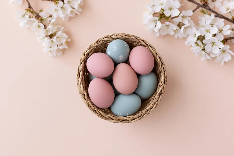 Pastel-painted easter eggs in a small woven basket with cherry blossoms on a soft peach background