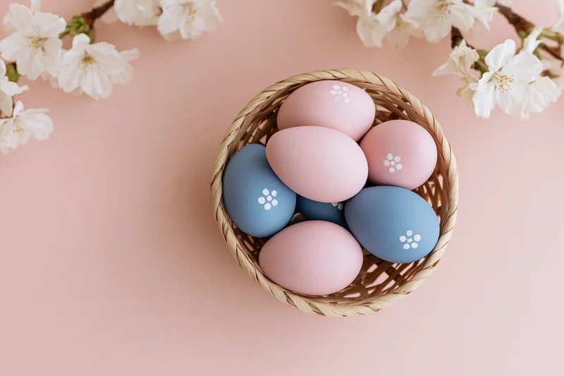 Pastel-painted easter eggs in a woven basket with cherry blossoms on a pink surface