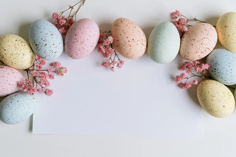 Pastel-speckled easter eggs and pink blossoms arranged in a flat lay around a blank white paper with copyspace