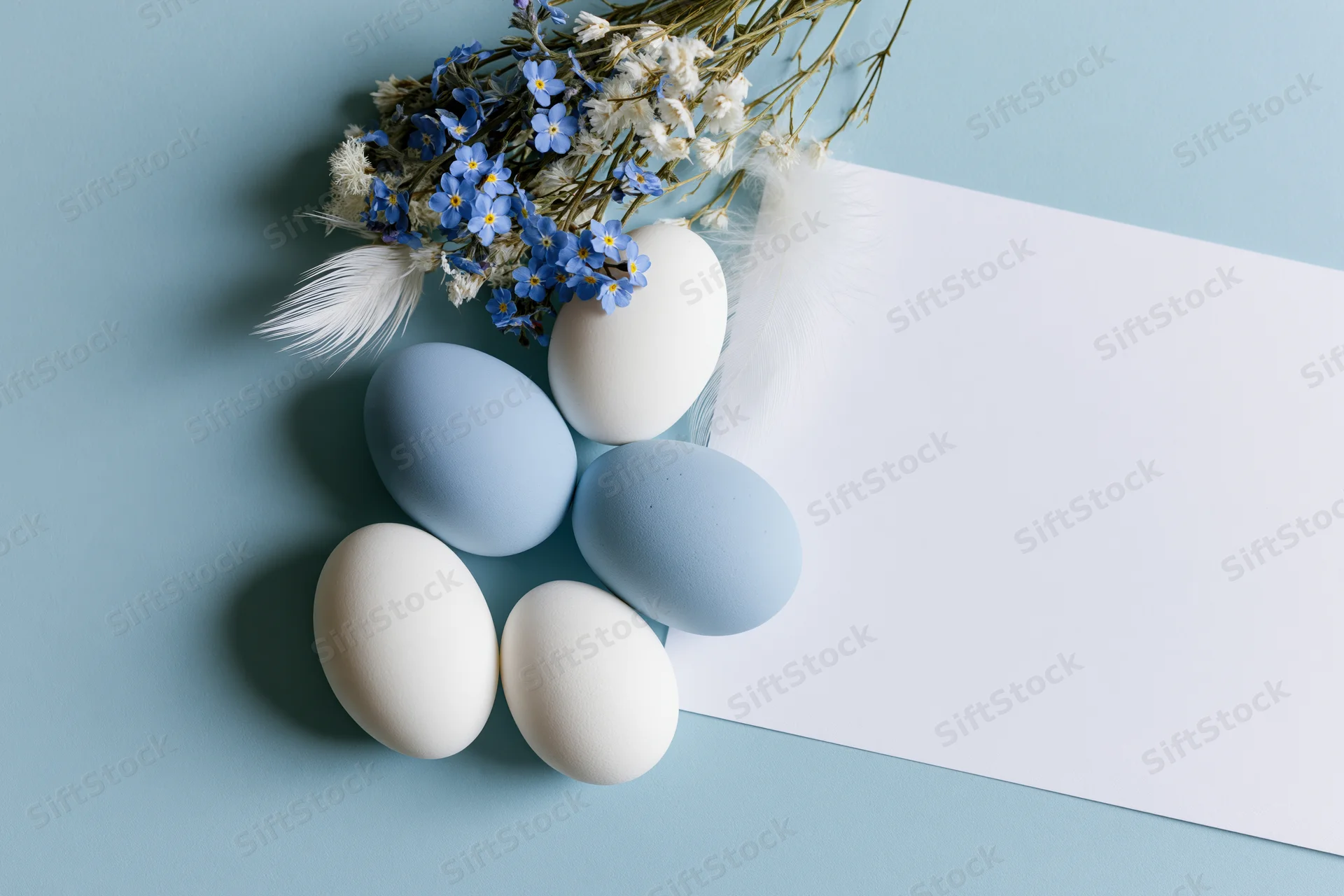 Pastel blue and white easter eggs arranged with dried forget-me-not flowers and feathers on a light blue surface beside a blank white card