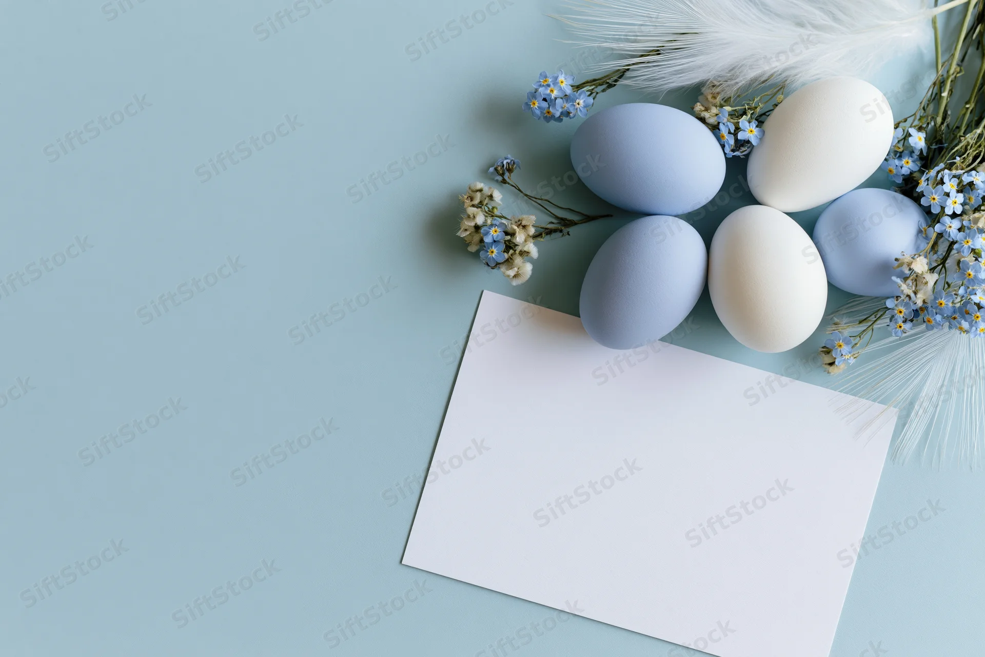 Pastel blue and white easter eggs arranged with forget-me-not flowers and feathers on a light blue surface beside a blank card