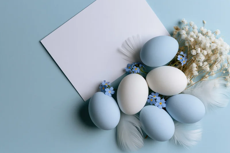 Pastel blue and white easter eggs arranged with forget-me-nots and feathers on a light blue surface beside a blank paper sheet