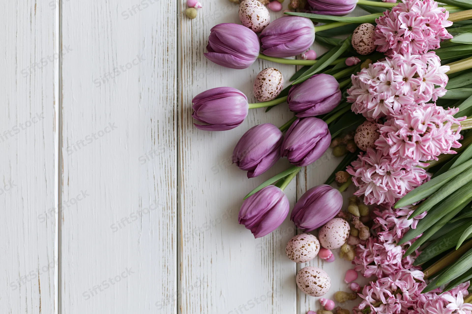 Purple tulips and pink hyacinths arranged with speckled easter eggs on a white wooden surface
