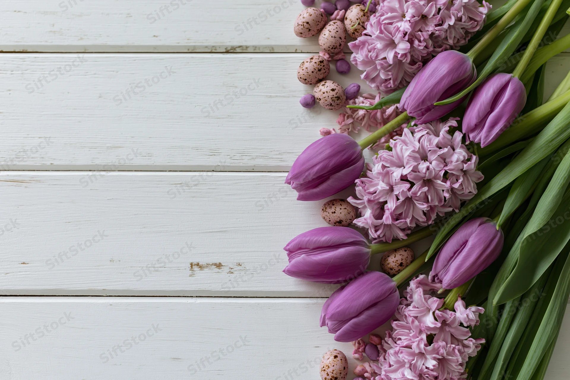 Purple tulips and pink hyacinths arranged with speckled easter eggs on white wooden surface