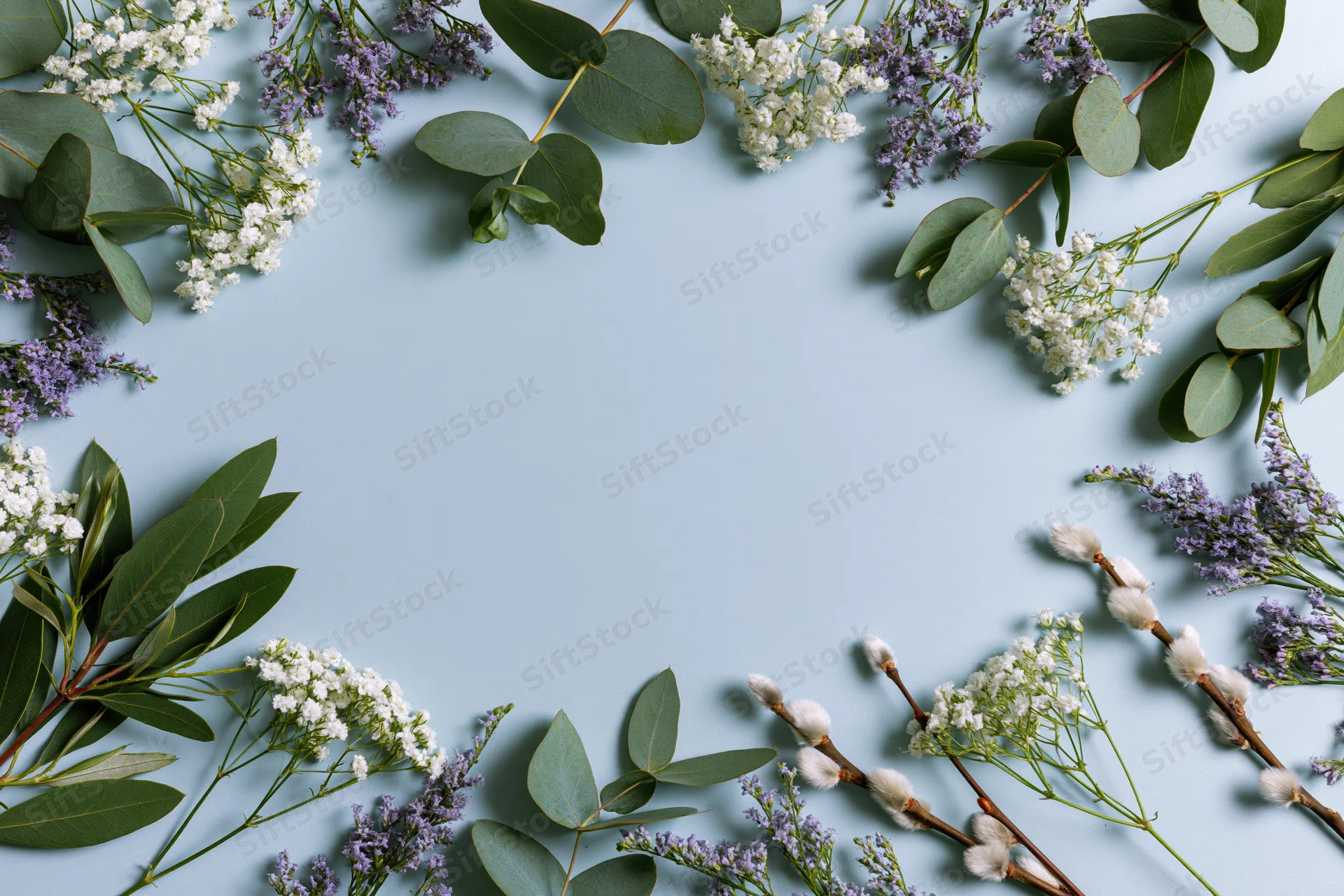 Scattered eucalyptus branches, small white gypsophila, purple lavender, and cotton buds arranged in a flat lay frame on a light blue background with copyspace