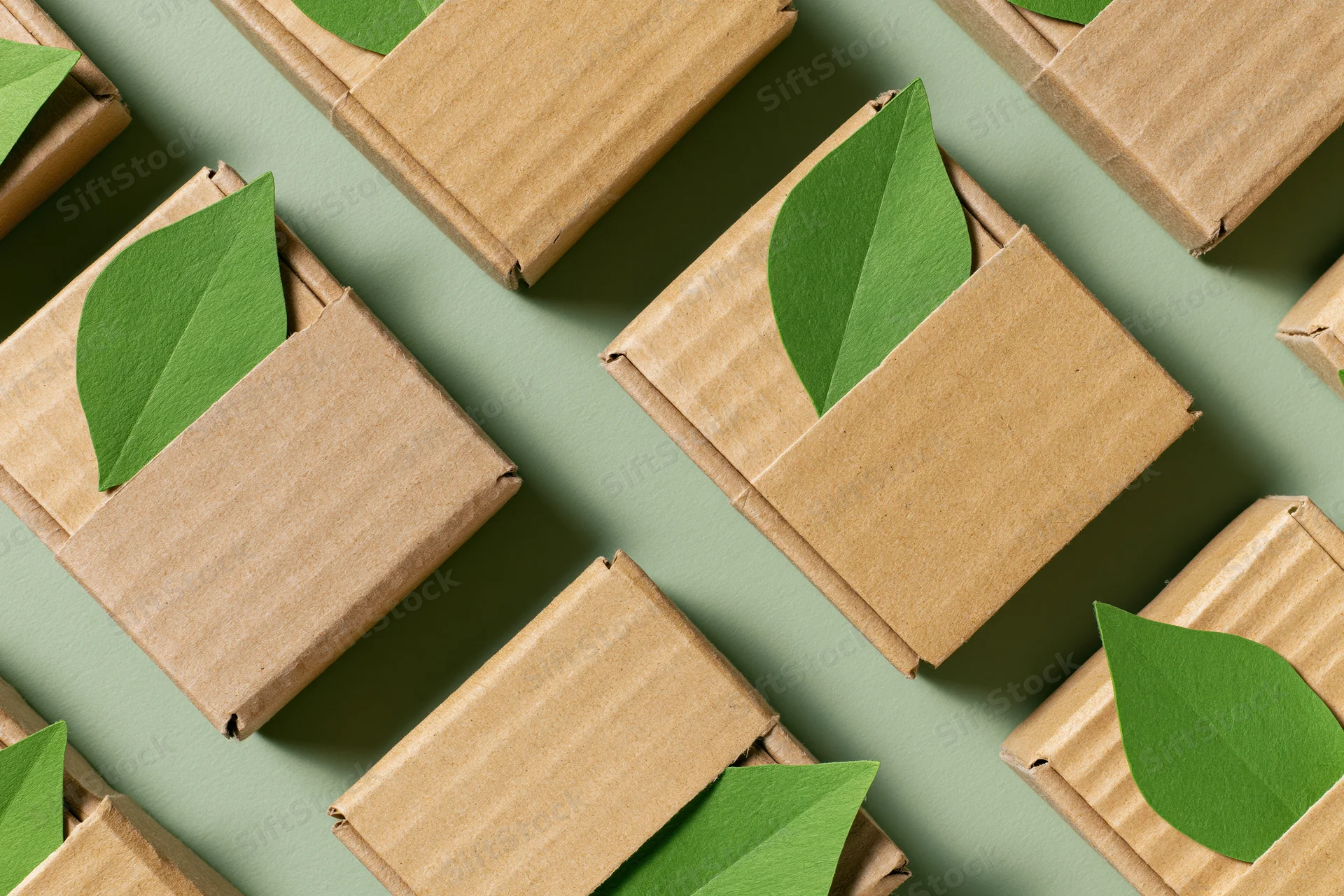 Small corrugated cardboard shipping boxes arranged in a flat lay pattern with green leaves peeking from flaps on a muted green surface