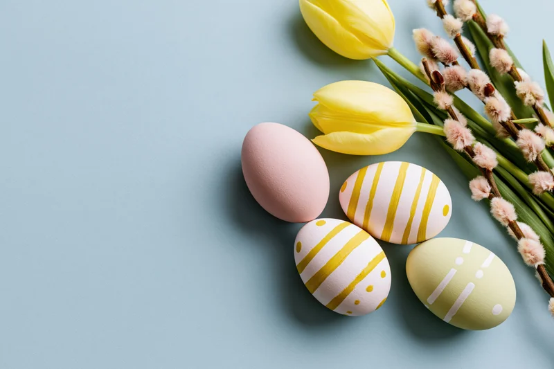 Hand-painted easter eggs and spring flowers arranged in flat lay on blue background