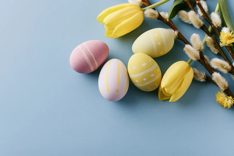 Hand-painted easter eggs and spring flowers arranged in flat lay on blue background