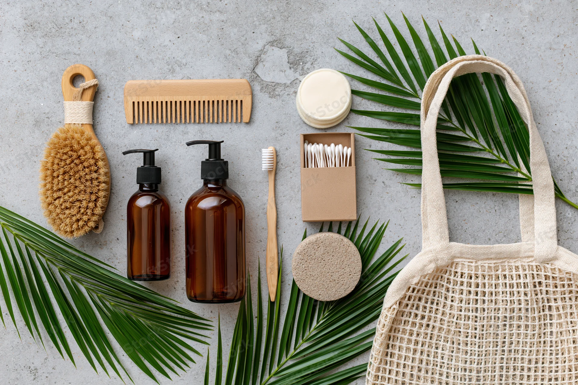 Zero-waste bathroom essentials arranged flat on concrete surface with palm leaves and mesh tote bag