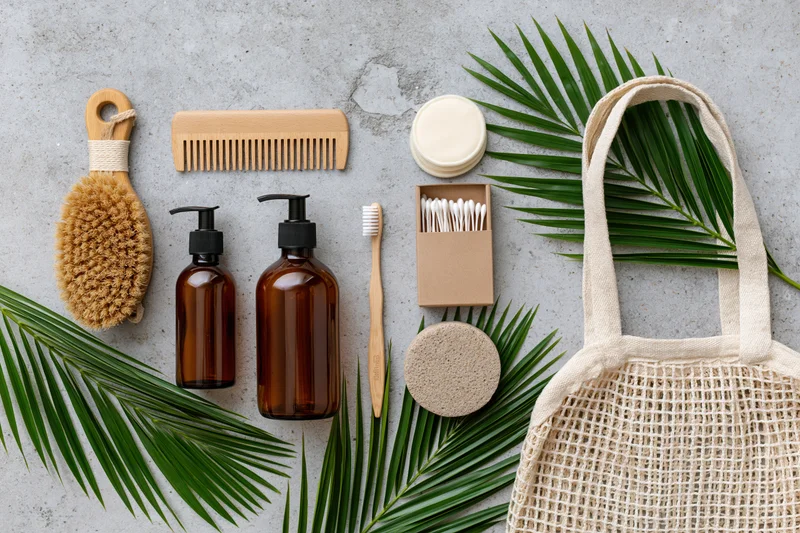Zero-waste bathroom essentials arranged flat on concrete surface with palm leaves and mesh tote bag