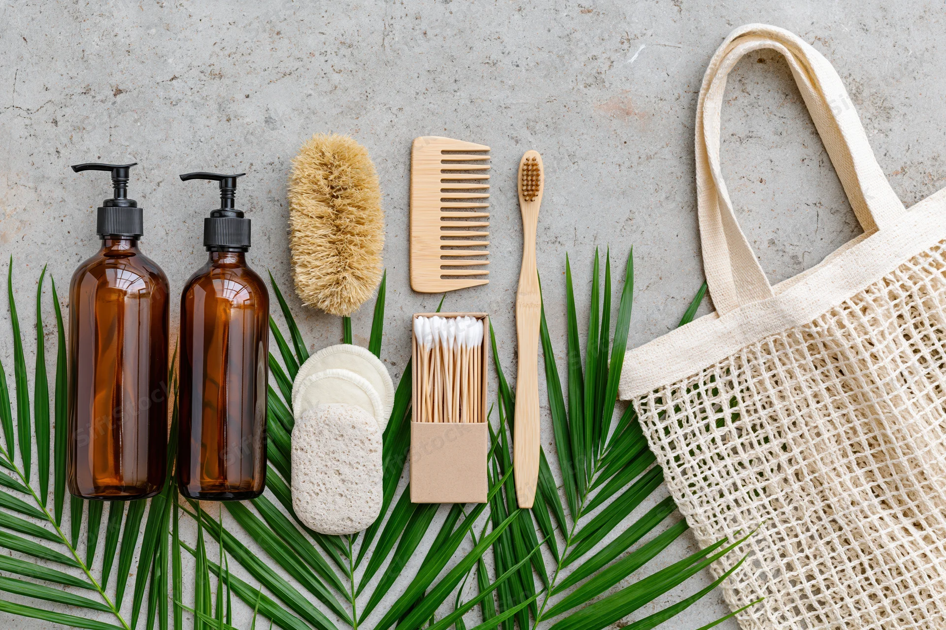 Zero-waste bathroom essentials arranged flat on concrete with palm leaves and a mesh tote bag
