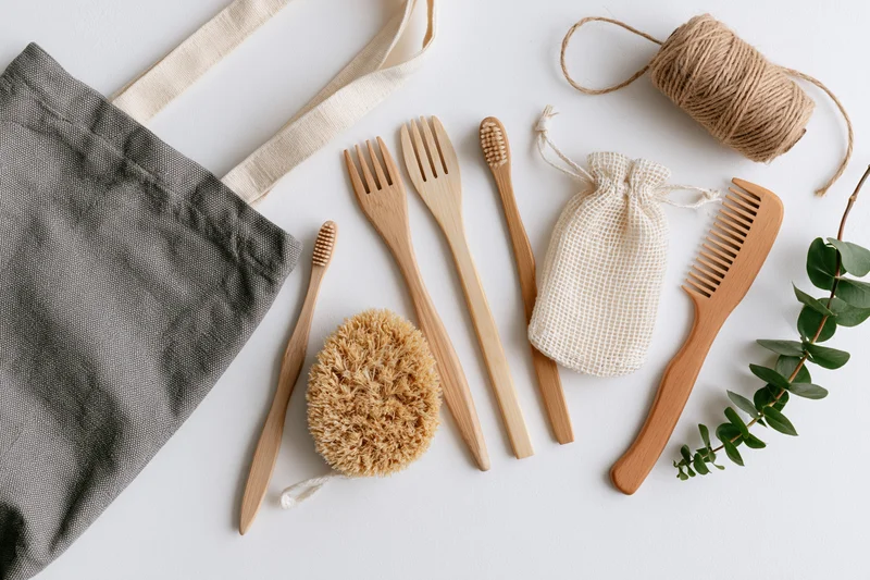 Zero-waste eco-friendly items arranged flat on white surface with bamboo toothbrushes, forks, comb, sponge, bag, twine, and eucalyptus leaf