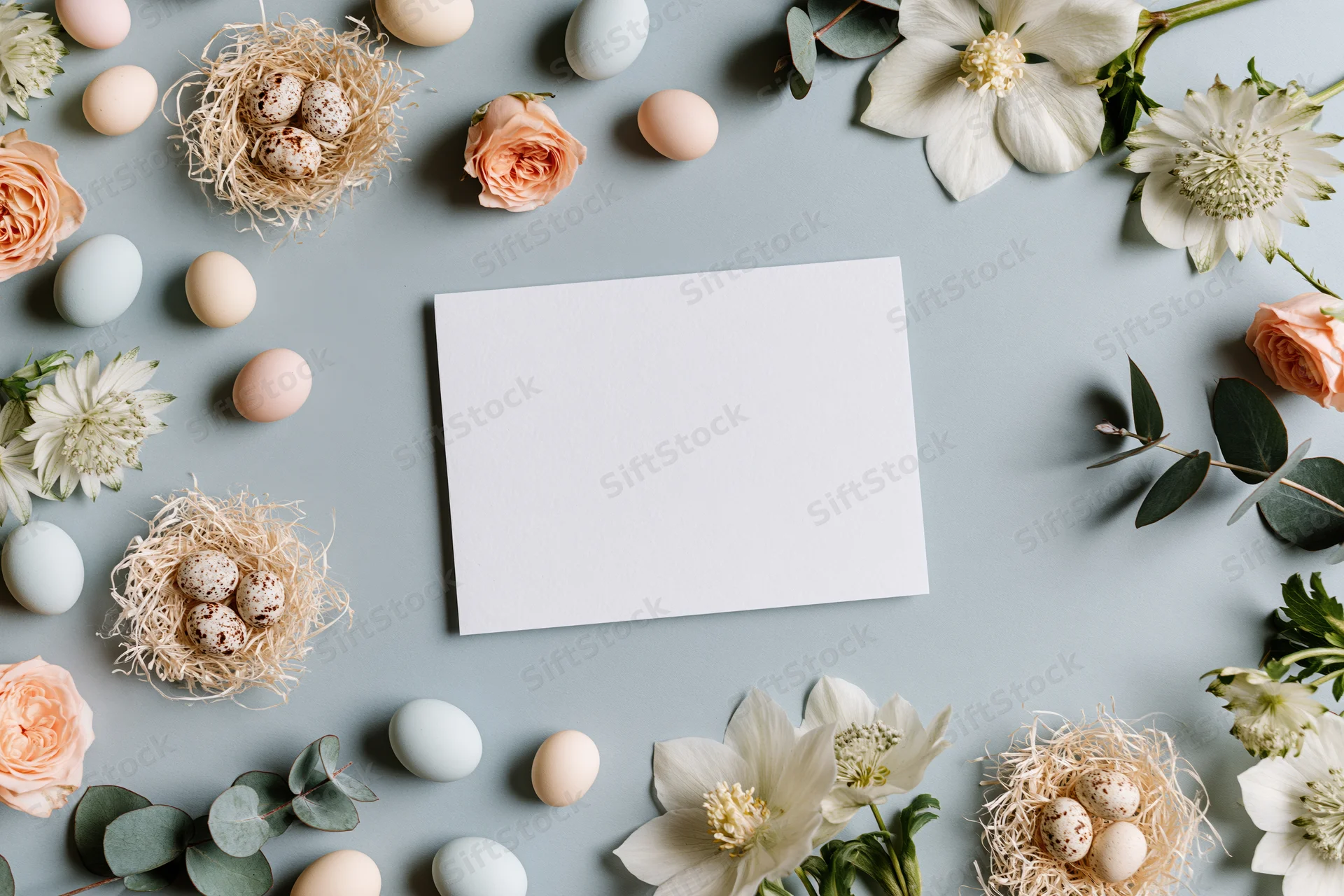 Blank white card centered on a flat lay with easter eggs, flowers, and nests arranged around it on a gray surface