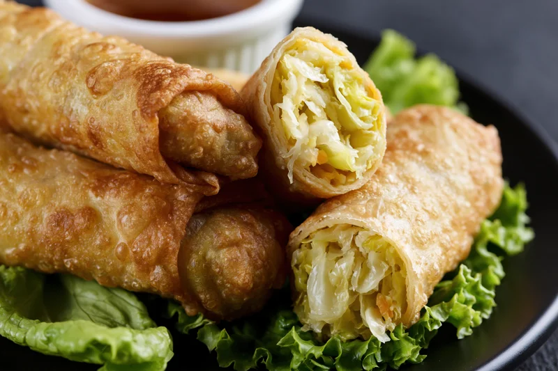 Four deep-fried egg rolls stacked on fresh green lettuce leaves on a black plate with dipping sauce in a white ramekin