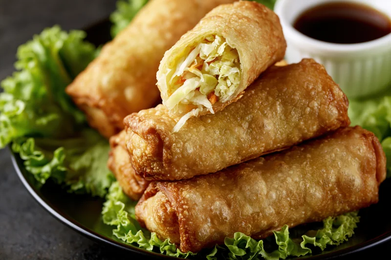 Four deep-fried egg rolls stacked on fresh green lettuce leaves with dipping sauce on a black plate