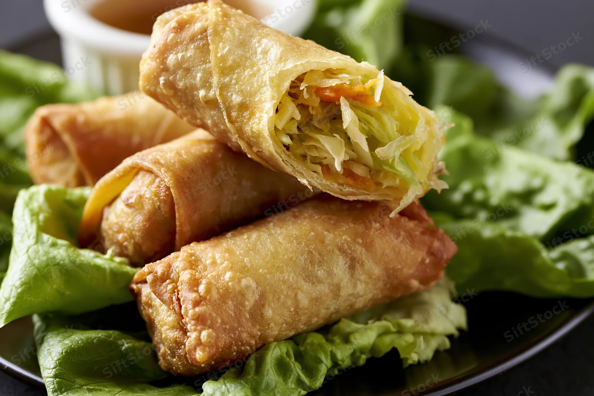 Four deep-fried egg rolls stacked on fresh green lettuce leaves on a black plate