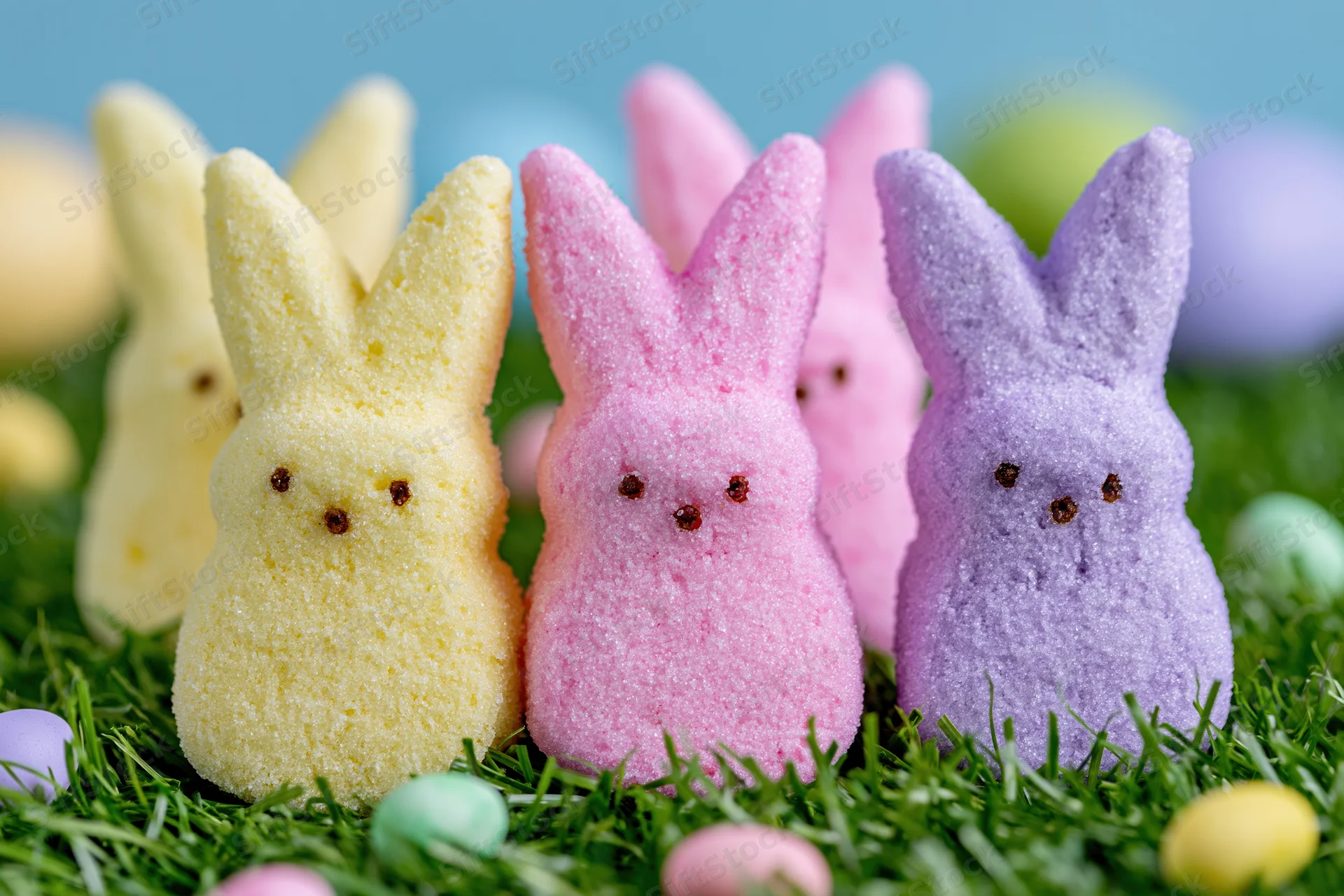 Marshmallow Peeps bunny candies in yellow, pink and purple arranged on green grass with small colorful eggs scattered around
