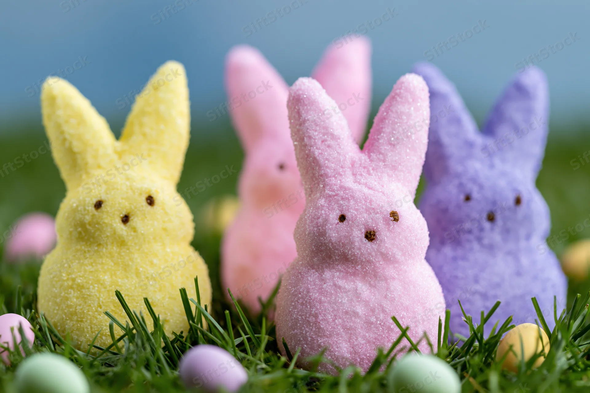 Marshmallow peeps bunny candies in yellow pink and purple arranged on grass with easter eggs around