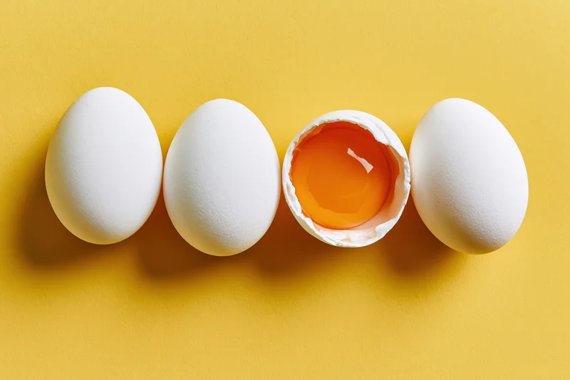 Four white chicken eggs in a row on a yellow surface with one cracked open revealing the yolk