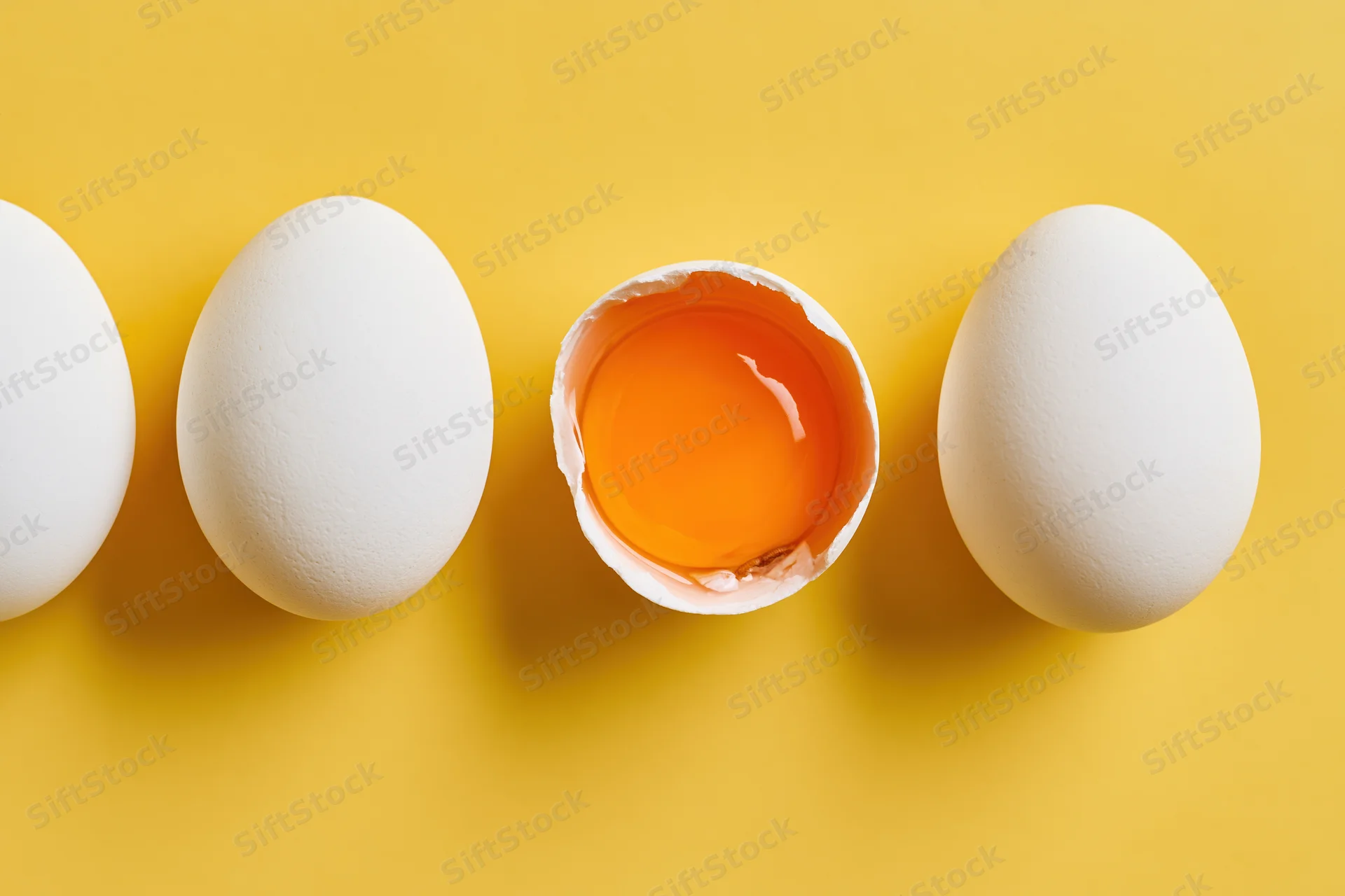 Four white chicken eggs in a row on yellow surface with one broken open showing yolk