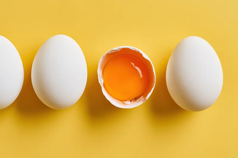 Four white chicken eggs in a row on yellow surface with one broken open showing yolk