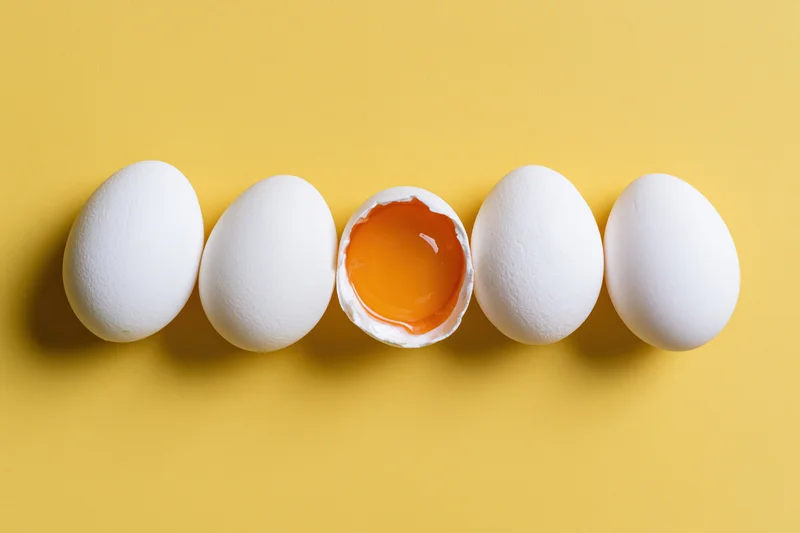 Five white chicken eggs in a horizontal row on a bright yellow surface with one cracked open to reveal the yolk