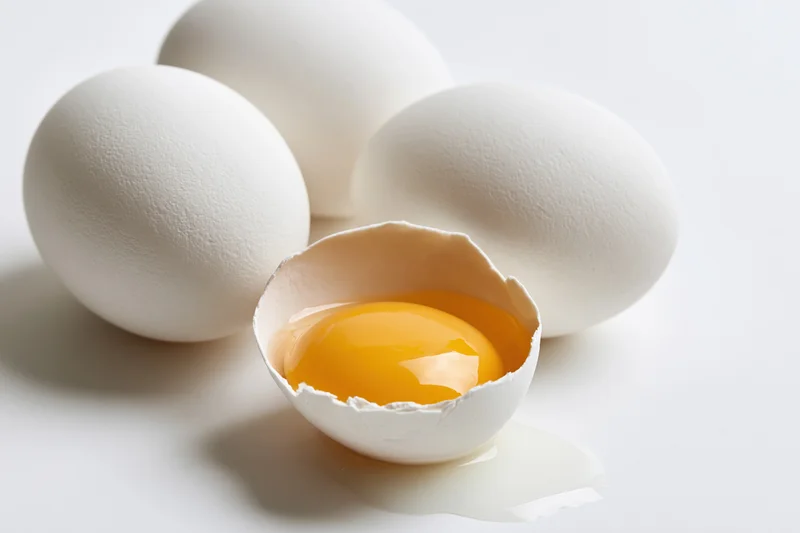 Four white chicken eggs on plain white surface one cracked with yolk visible