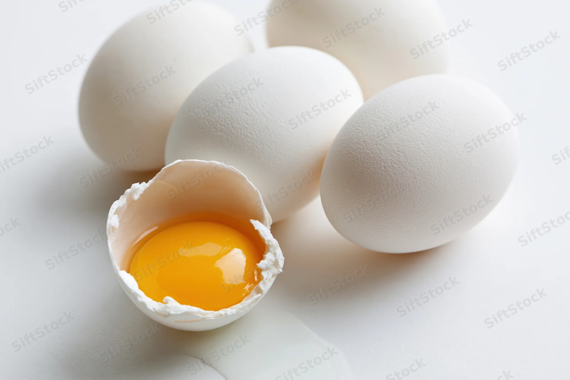 Cracked chicken egg with yolk visible on plain white surface alongside whole eggs