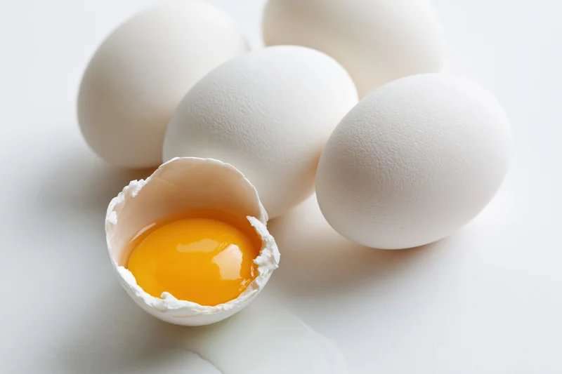 Cracked chicken egg with yolk visible on plain white surface alongside whole eggs