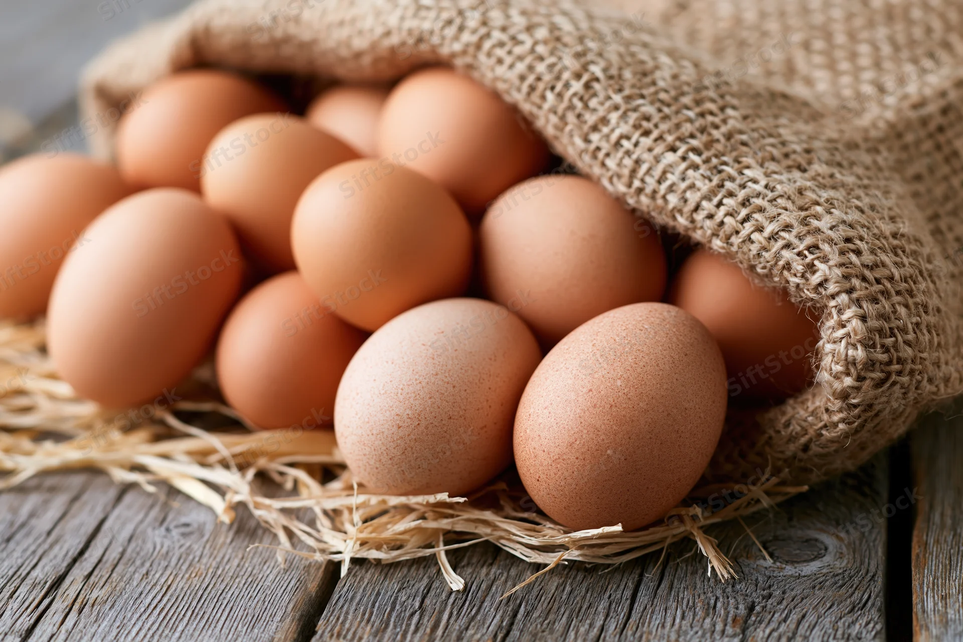Brown chicken eggs spilling from open burlap sack onto straw on wooden table