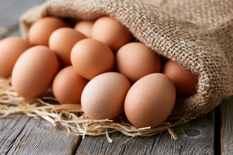 Brown chicken eggs spilling from open burlap sack onto straw on wooden table